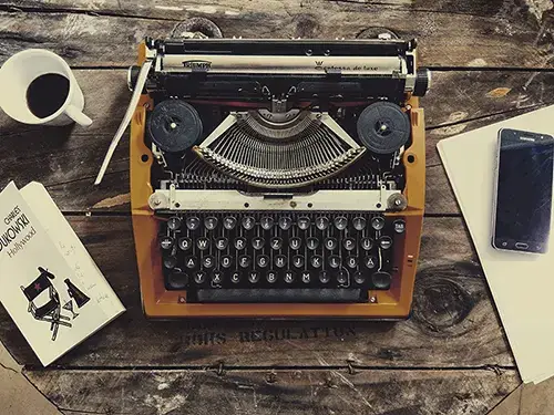 Vintage-Schreibmaschine, Bukowski-Buch, Kaffee und Smartphone auf Holztisch. Kontrast: Old School Texten vs. moderne SEO-Texte.