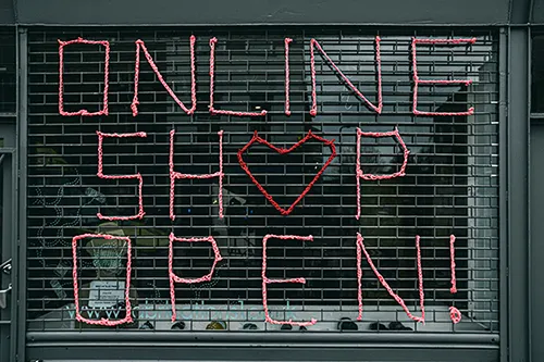 Dunkles Rollgitter eines Ladengeschäfts, in das mit farbiger Schnur der Schriftzug "ONLINE SHOP OPEN!" eingeflochten ist. Das O in Shop ist als rotes Herz gestaltet.