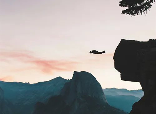 Symbolbild: Silhouette einer Person, die bei Dämmerung einen weiten Sprung zwischen zwei steilen Felsklippen wagt.