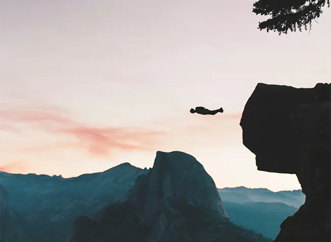 Symbolbild: Silhouette einer Person, die bei Dämmerung einen weiten Sprung zwischen zwei steilen Felsklippen wagt.